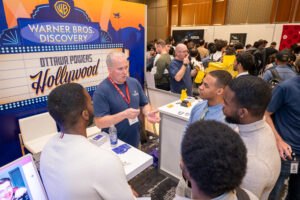 Jobseekers connect with Warner Bros Discover at the Discover Technata Career Fair 2024. Andrew Emmons is pictured chatting with four attendees, in front of Warner Bros' Hollywood-esque setup.