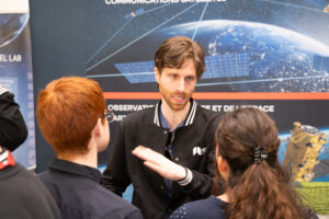 MDA Space, winners of the Best in Tech award, chatting with two engaged jobseekers. Behind the MDA representative is their exhibitor display showing a satellite above earth.