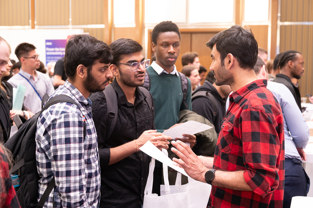 Four jobseekers chat with each other at the 2024 Discover Technata Career Fair. One of them is holding his resume and a Hub350 swag tote bag.