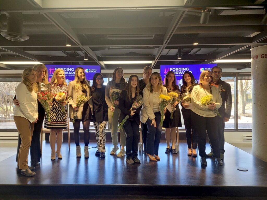 Members of the Kanata North business and tech community pose on the Hub350 stage for a photo, holding flowers and everyone smiling. MP Jenna Sudds is front and center.