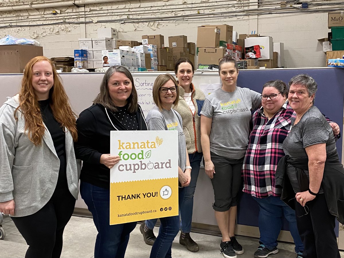 Cara-Leigh Wyllie stands holding the Kanata Food Cupboard sign, amongst a team of volunteers in the KFC's old warehouse at 340 Legget Drive.