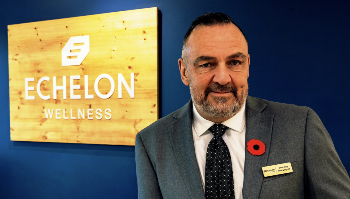 Headshot of Jean-Guy Bourguignon, Vice-President of Operations at Echelon Wellness. Jean-Guy smiles in a suit adorned with a red poppy, in front of the 'Echelon Wellness' location in Kanata, Ontario.