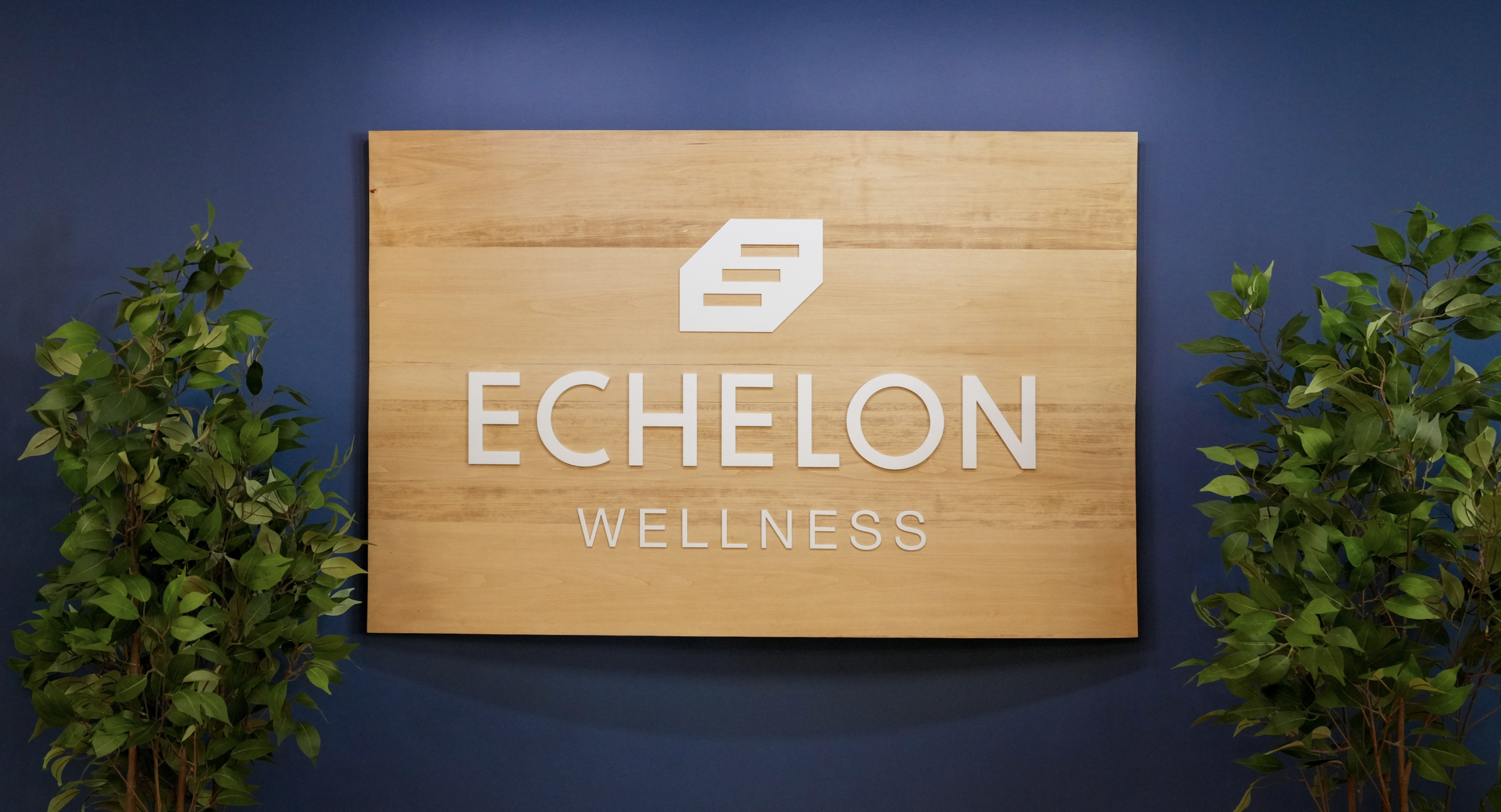The Echelon Wellness logo on a wooden background surrounded by foliage; displayed at the entrance of Echelon Wellness' Kanata location.