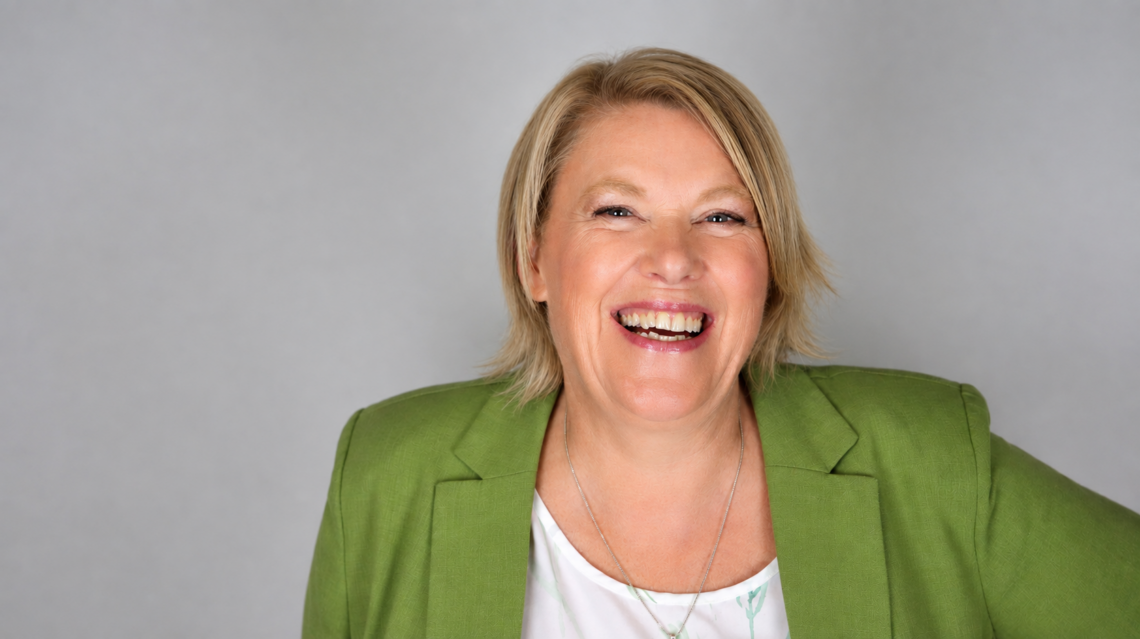 Headshot of Lisa Hewston in a green blazer on grey background. She is laughing.